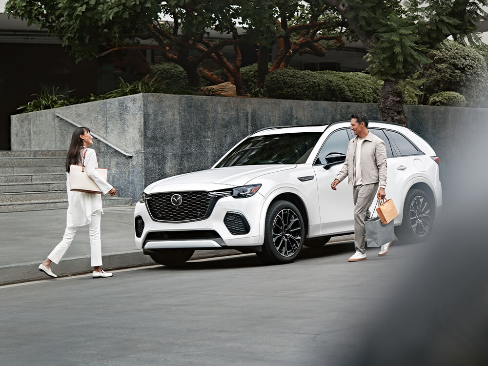 Deux personnes marchent à côté d’un CX-70 blanc stationné en bordure de rue.