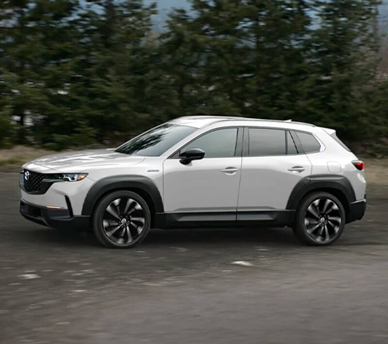 A white CX-50 Hybrid drives alongside trees