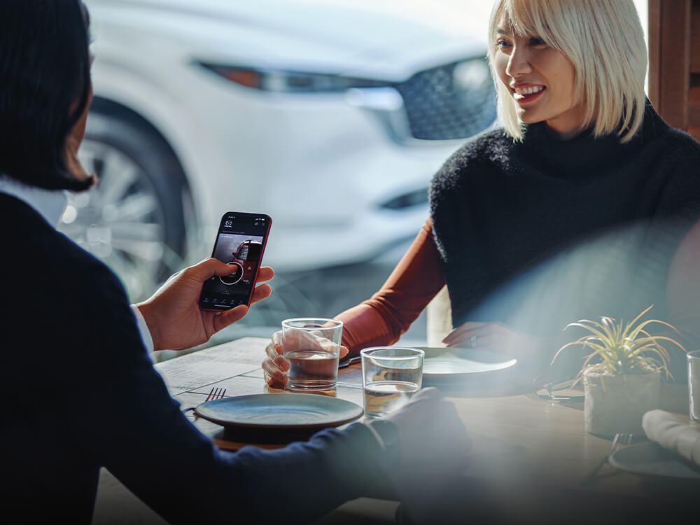Une femme assise à une table tient un téléphone avec l’appli MyMazda ouverte.