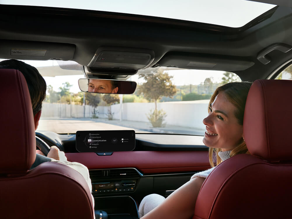 Depuis le siège arrière, vue vers l’avant à bord d’un Mazda CX-70 hybride léger avec un élégant intérieur en cuir Nappa rouge grenat. Une femme sur le siège passager se tourne pour regarder en arrière. Un homme conduit, ses yeux sont visibles dans le rétroviseur. 