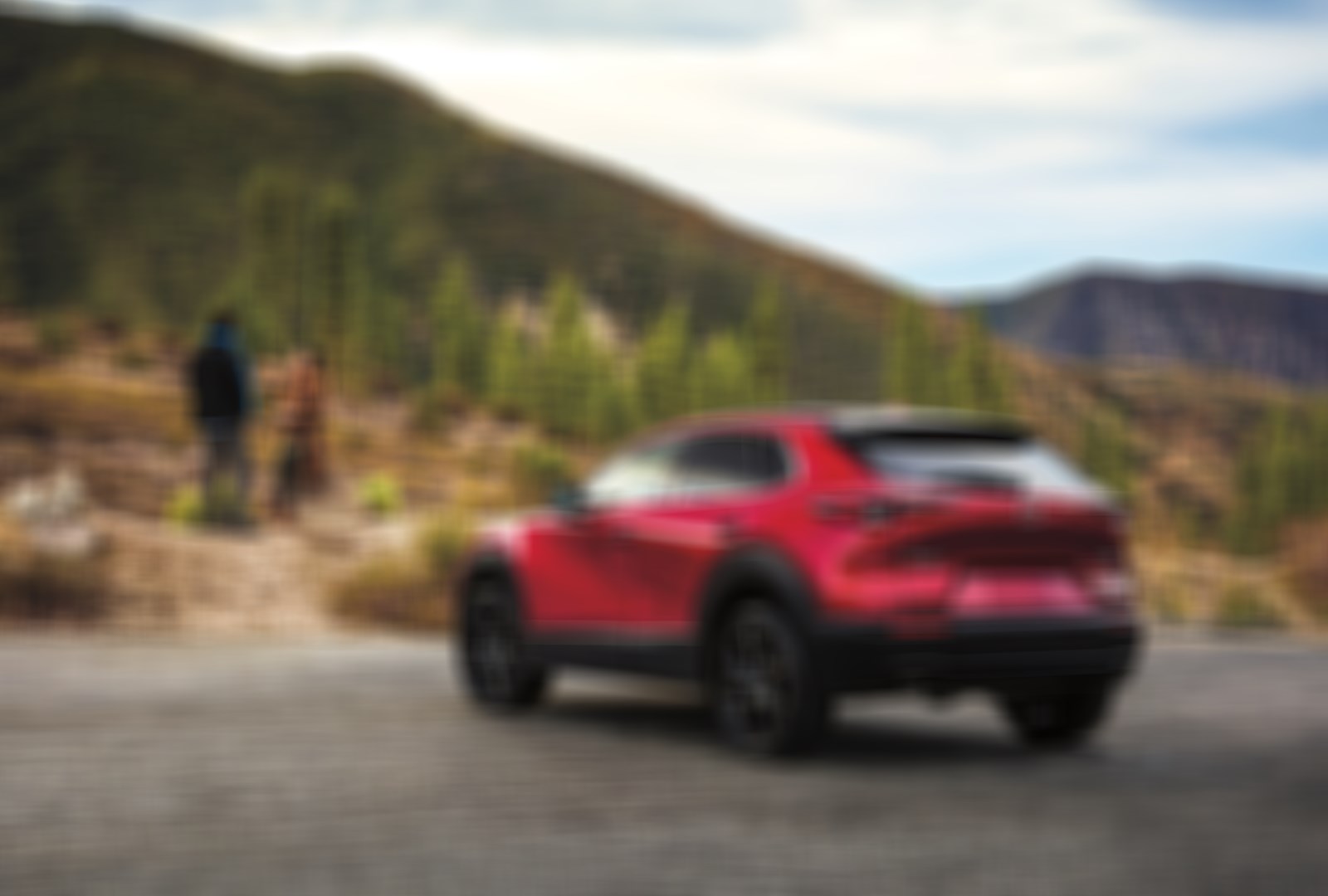 Soul Red Crystal Metallic CX-30 parked below rolling hills as a couple converses in the background.