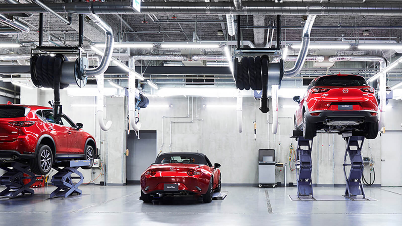 Three red Mazdas are being worked on in a garage. Two are parked and one is elevated. 