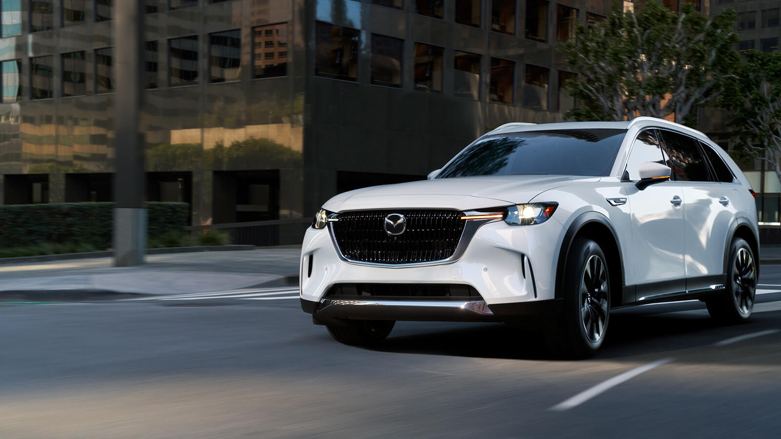 A white Mazda is in front of a crosswalk on an urban street with buildings in the background.  