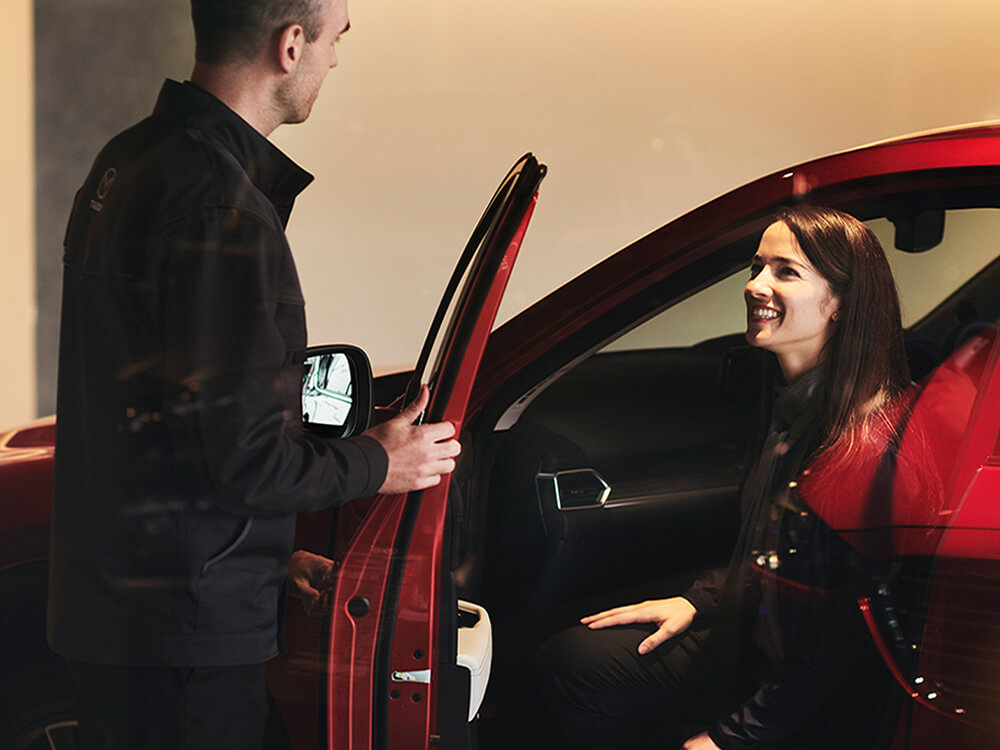 A man opens the door to a red Mazda. A woman sitting inside the car is smiling at him. 