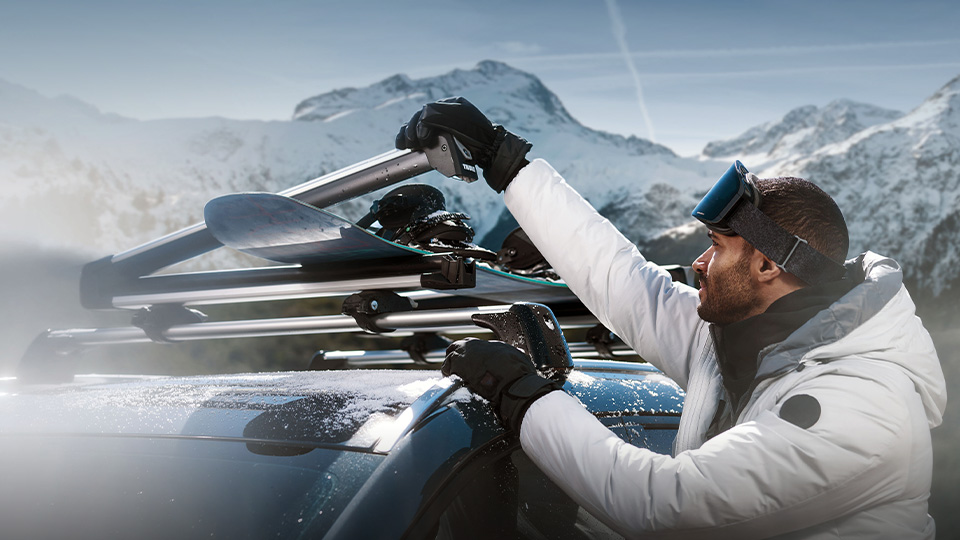 Man dressed for skiing holding a ski beside his Mazda CX-50 on top of a snowy mountain.