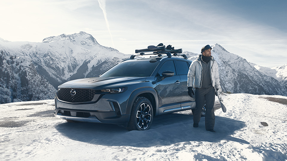 Man dressed for skiing holding a ski beside his Mazda CX-50 on top of a snowy mountain.
