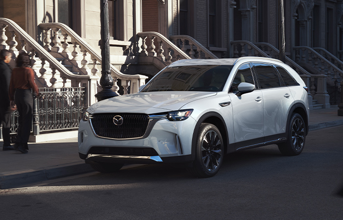 Mazda CX-90 parked on city street, morning light shines on the apartments in the background.  
