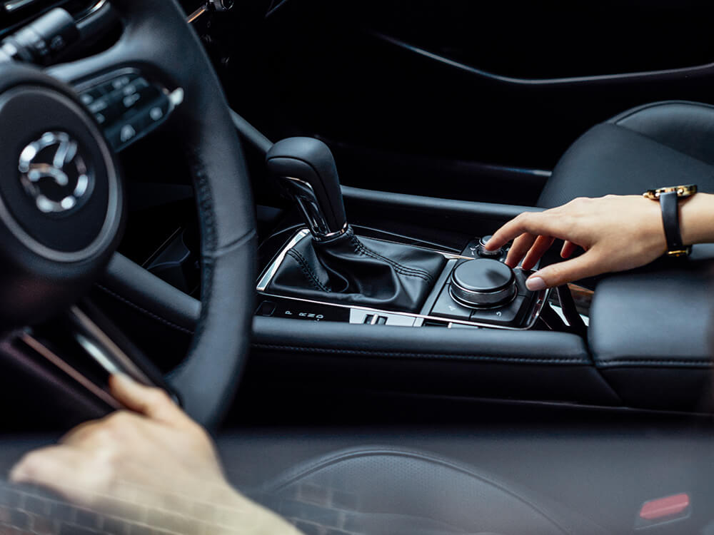 Un conducteur dans l’habitacle d’une berline Mazda3, une main tendue vers le bouton de la console centrale HMI. 