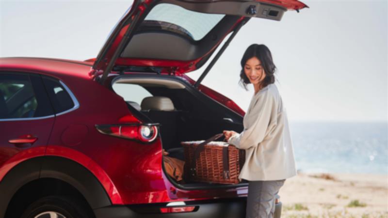 Soul Red Crystal Metallic CX-30 parked lakeside. The rear liftgate is open, and a woman is removing a wicker picnic basket/beach box from the cargo hold.