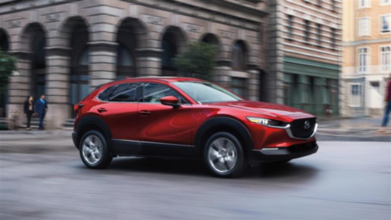 7/8 profile close up shot of Soul Red Crystal Metallic Mazda CX-30 driving through city, stately stone buildings in background. 