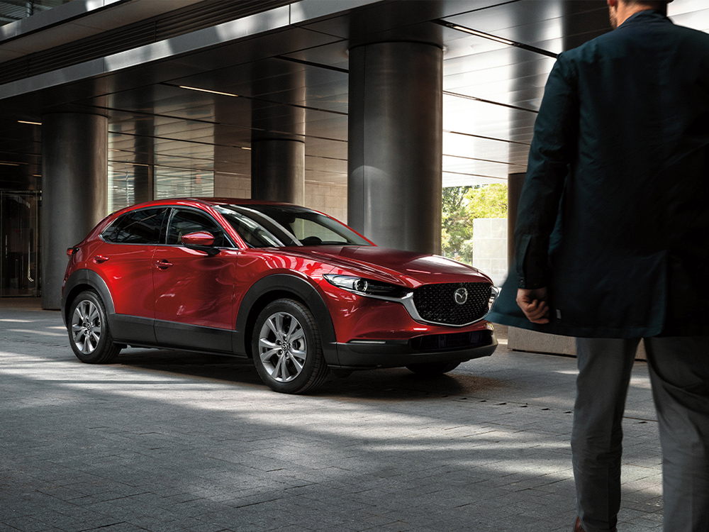 Un homme au premier plan s’approche d’un Mazda CX-30 rouge vibrant cristal métallisé garé de ¾ face dans l’allée d’un bâtiment moderne.