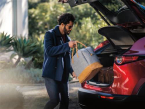 A man is placing a bag into the cargo hold of Soul Red Crystal Metallic CX-30 with liftgate open. Other bags are visible in the cargo hold. 