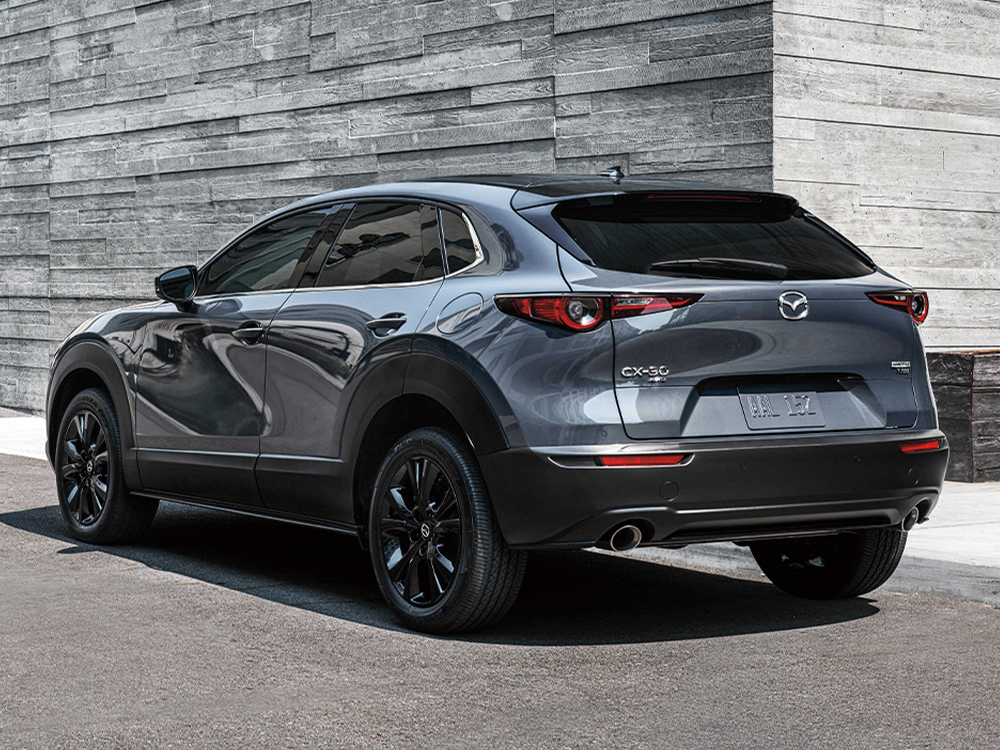 Profil de ¾ arrière d’un Mazda CX-30 gris mécanique métallisé garé devant des murs en pierre grise élégants et minimalistes.