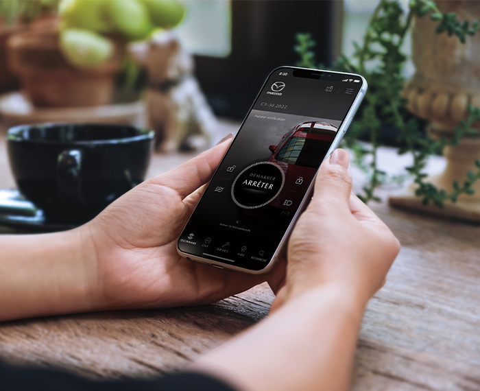 Des mains tenant un téléphone intelligent qui affiche l’écran d’accueil de l’application MyMazda.