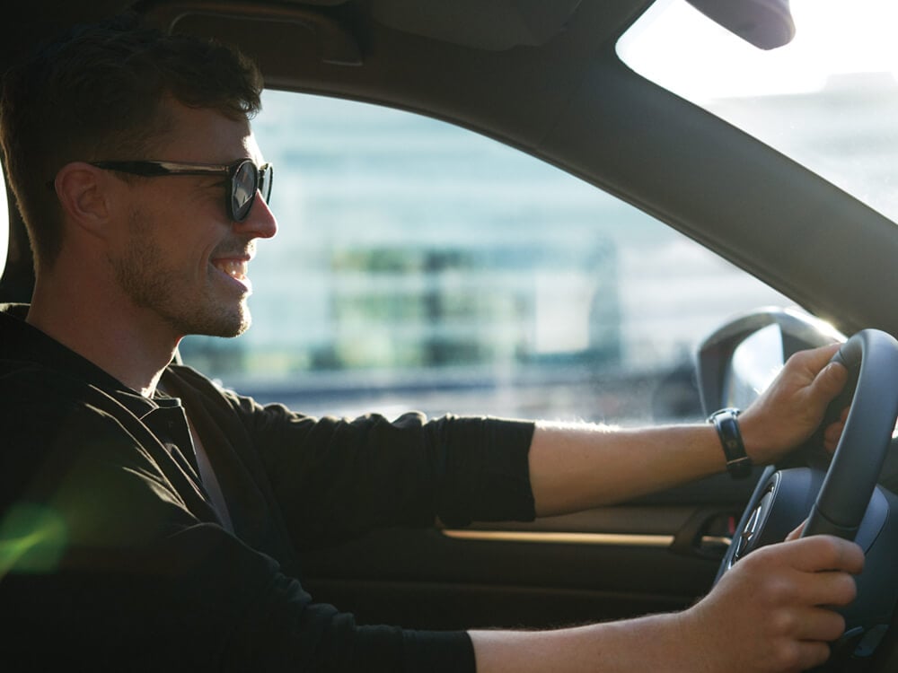 Vue rapprochée depuis le siège passager d’un homme souriant portant des lunettes de soleil au volant d’un CX-5.