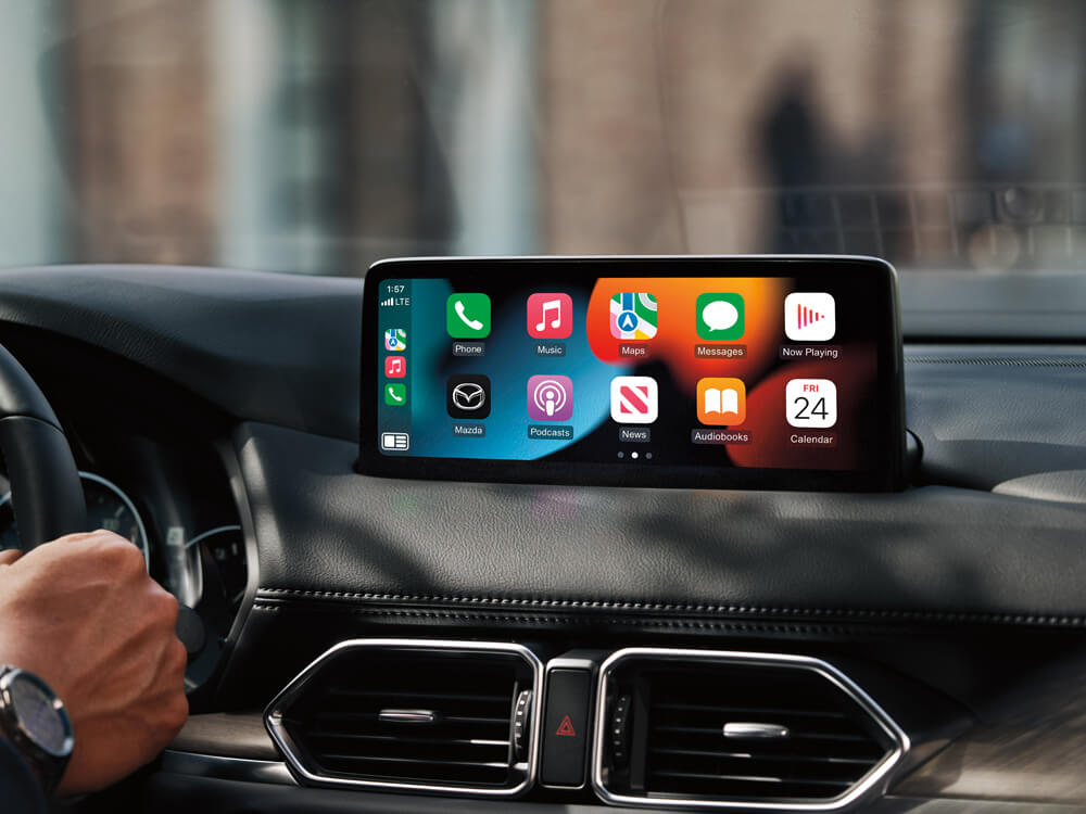 Close up of dashboard vents and screen display showing app icons. To the left, the driver’s hand holds the steering wheel.