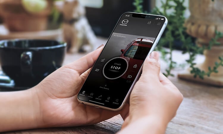 Two hands holding smartphone with MyMazda app activated on screen, engine start/stop button displayed. 