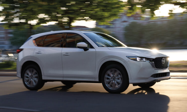 Plan de profil d’un CX-5 blanc rhodium qui circule avec des bâtiments majestueux et des arbres flous en arrière-plan. 