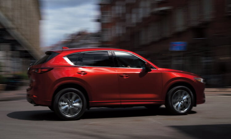 Soul Red Crystal Metallic   CX-5 driving through city’s main street intersection on a sunny day, buildings are motion blurred. 