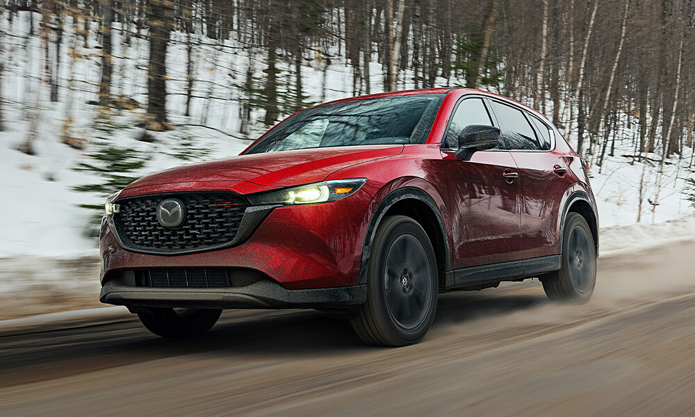 Vue latérale avant d’un Mazda CX-5 rouge vibrant cristal qui roule avec une forêt enneigée floue en arrière-plan.