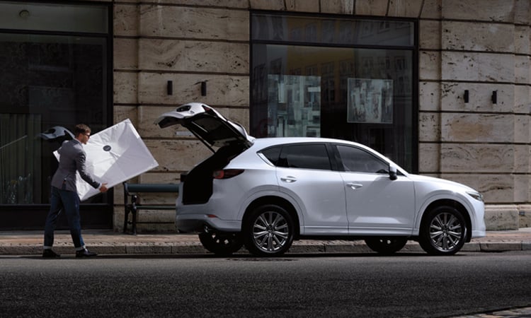 Man carries white frame to Rhodium White Metallic Mazda CX-5 parked on city street with liftgate open. 