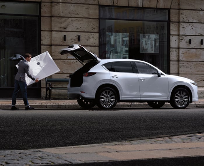 Un homme transporte un cadre blanc vers un Mazda CX-5 blanc rhodium métallisé garé dans la ville avec le hayon ouvert. 