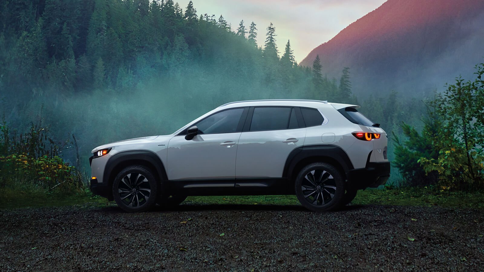 White Mazda CX-50 Hybrid parked in profile on dirt in clearing in forest. In background are trees and mountains.