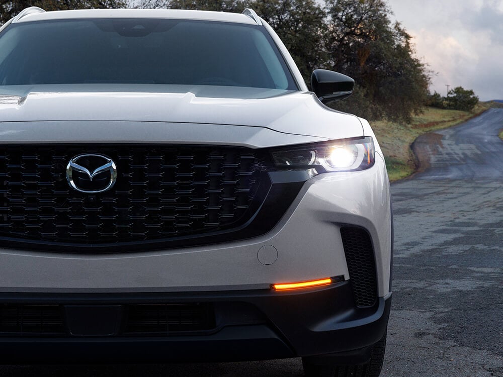 Close up low shot of Mazda CX-50 Hybrid grill and driver’s side headlamp. Rural road and trees in background.