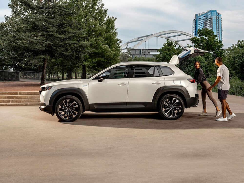 Profile view of CX-50 Hybrid with liftgate open, a man and a woman are at the rear, the woman is reaching in the cargo hold.