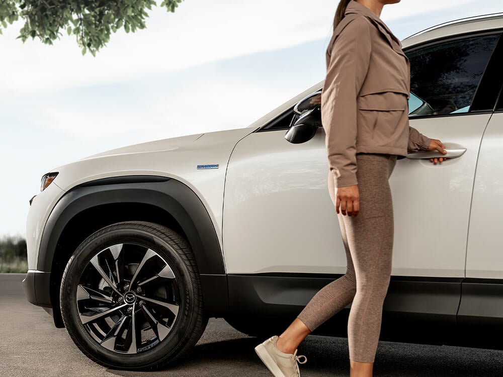 From low, a woman in athletic clothing is opening the driver’s door of Mazda CX-50 Hybrid.