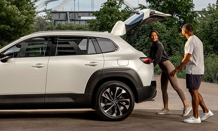 Profile view of CX-50 Hybrid with liftgate open, a man and a woman are at the rear, the woman is reaching in the cargo hold.