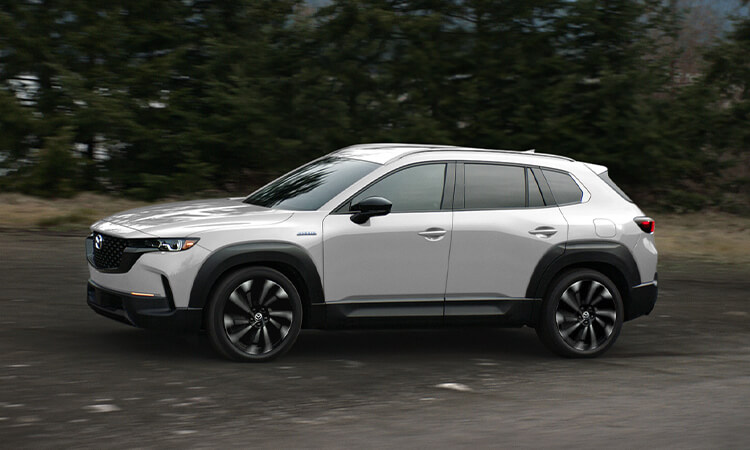 Wind Chill Pearl Mazda CX-50 Hybrid driving in profile close up, trees in background.