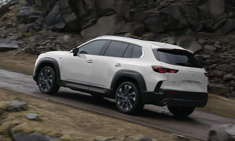 Un Mazda CX-50 hybride vent polaire nacré au 3/4 arrière escaladant une route de montagne pavée.