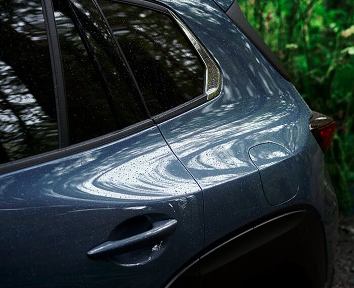 Close up of rear driver’s side panel of Ingot Blue Metallic Mazda CX-50, dense forest in background.