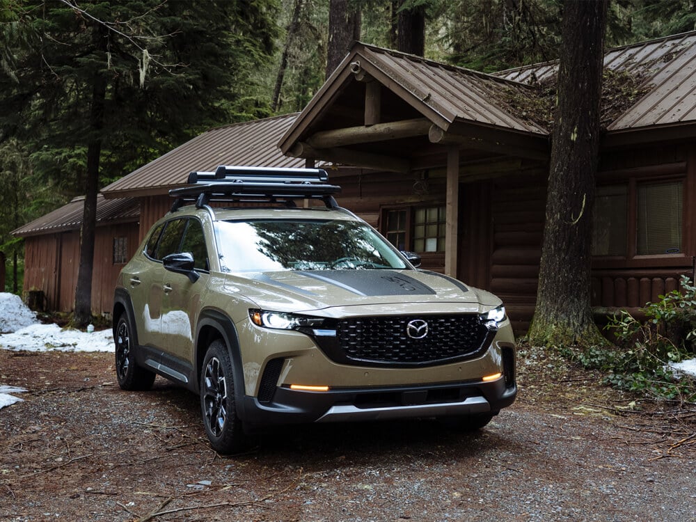 Vue de ¾ d’un CX-50 Meridian sable de zircon métallisé garé au 7/8 face devant une cabane en rondins dans la forêt.   .