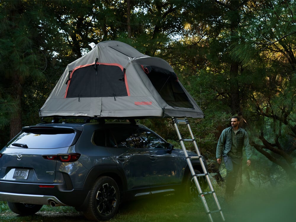 Un CX-50 bleu lingot métallisé avec une tente et une échelle installés sur le toit, garé sur la terre et l’herbe dans la forêt. Un amateur de plein air s’éloigne du véhicule. 