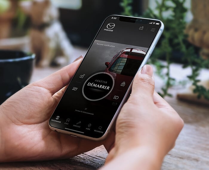 Deux mains tenant un téléphone avec l’appli MyMazda à l’écran affichant le bouton de démarrage/arrêt du moteur.  