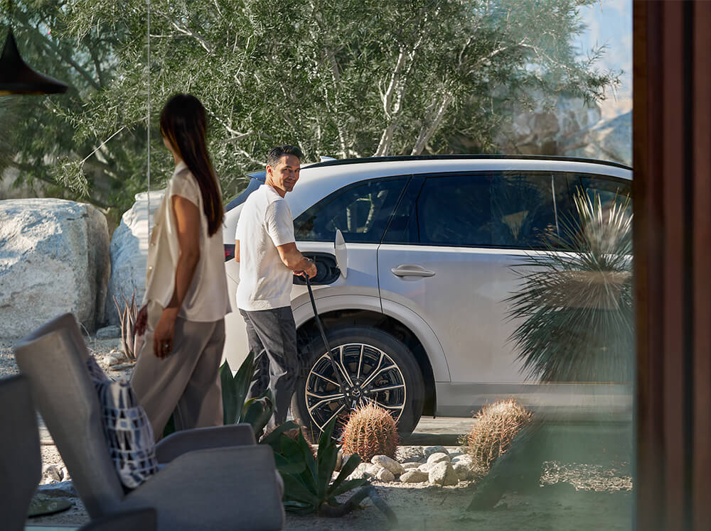 Couple smiles at each other in front of their modern home as a driver plugs his CX-70 PHEV in to charge.