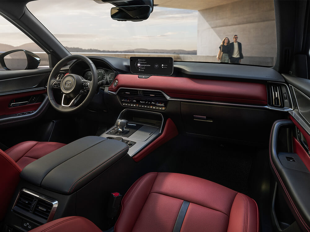 Shot from the passenger’s side inside the CX-70, peering out onto verdant landscape a couple returns to the vehicle as the sun sets.