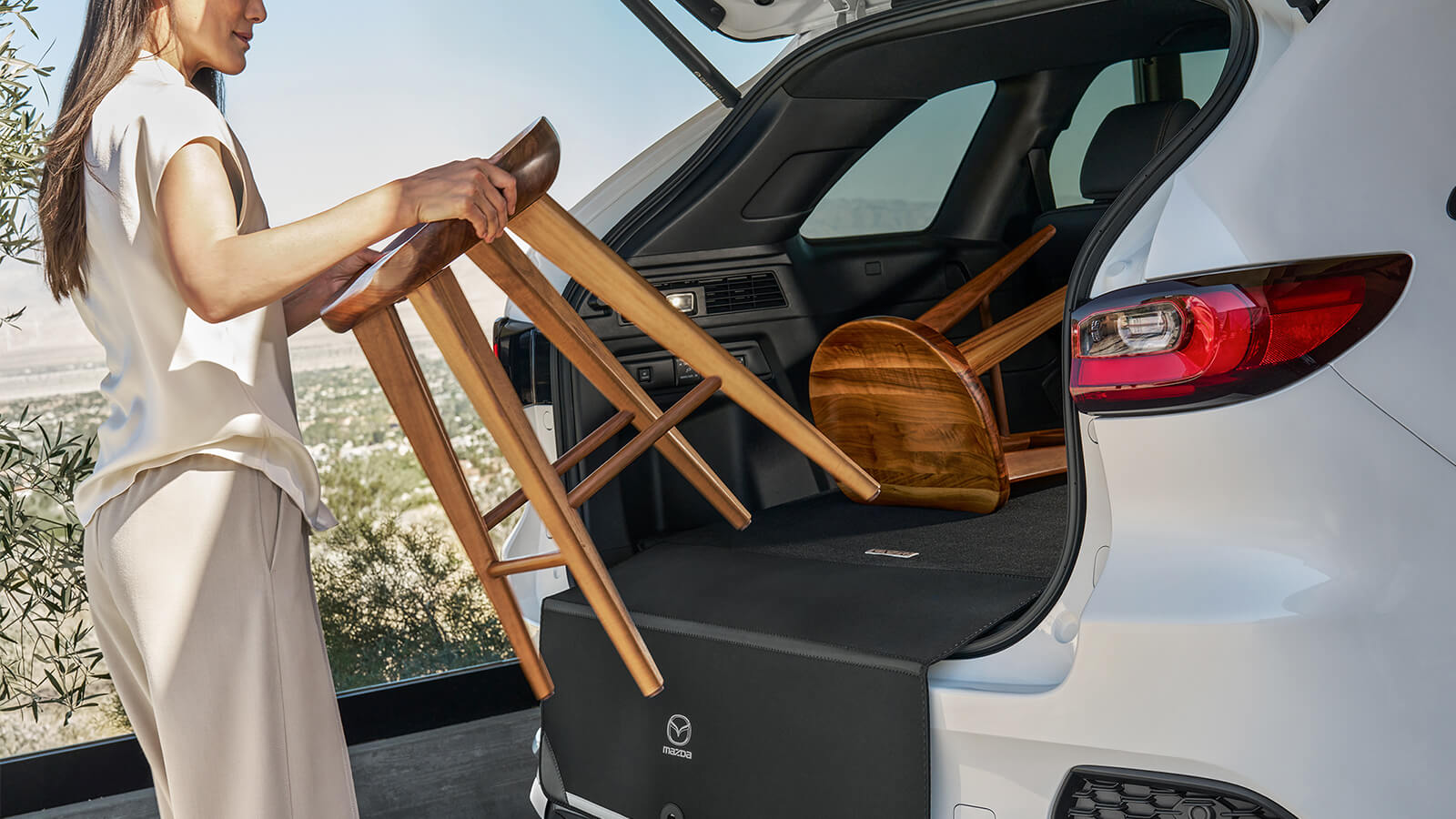 Behind the open lift gate of the CX-70 PHEV, a matt is draped over the liftgate’s edge as a driver lifts a wooden stool into the cargo area.