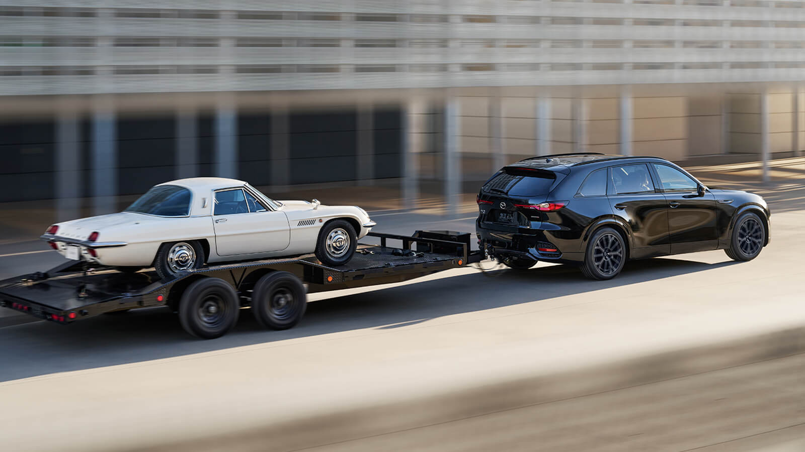 A CX-70 MHEV tows a classic, white Mazda sportscar on a vehicle trailer in the morning sun.