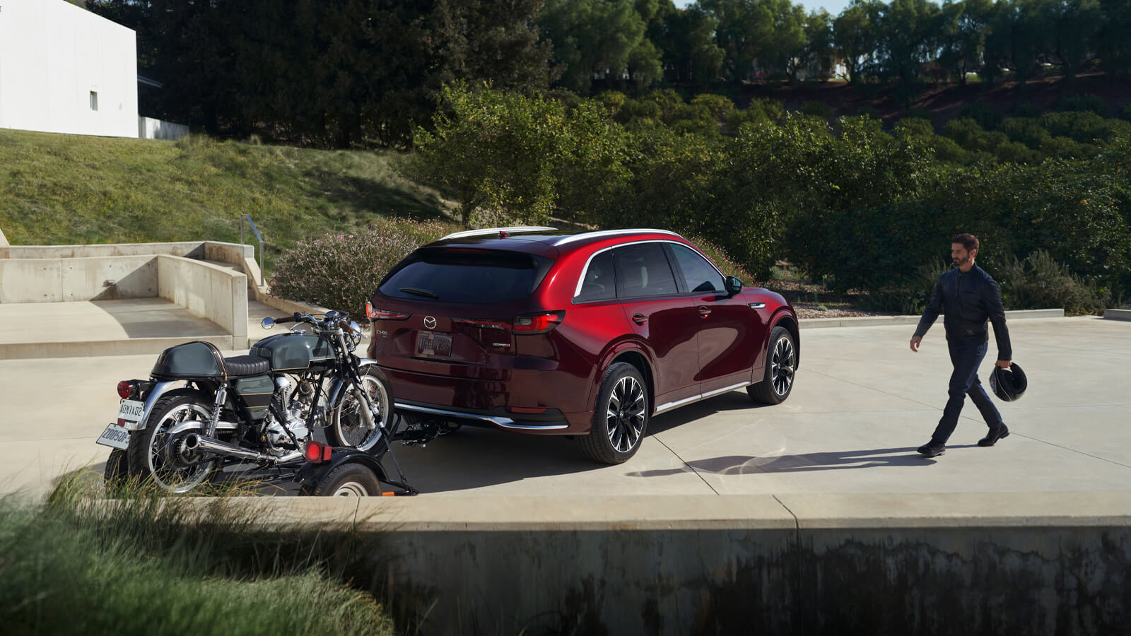 Artisan Red CX-90 is parked with a trailer on which is a motorcycle. A man in riding gear holding a motorcycle helmet walks toward the trailer.