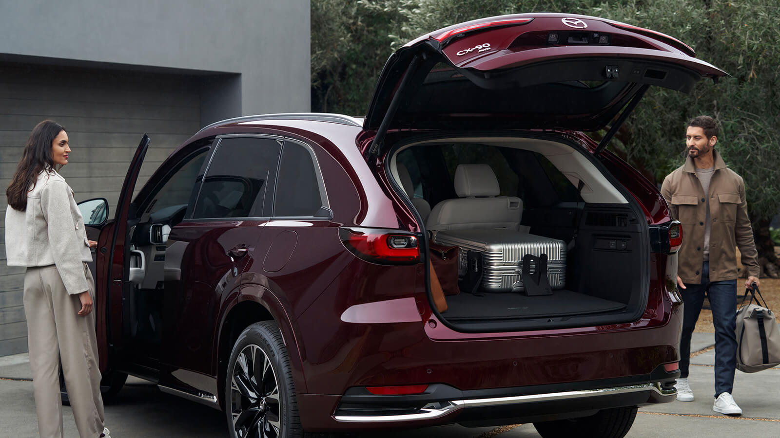 Un couple en train de charger un CX-90 rouge artisan; le hayon ouvert révèle une grande valise de couleur argent. L’homme se dirige vers l’arrière avec un sac, la femme ouvre la portière du côté conducteur.