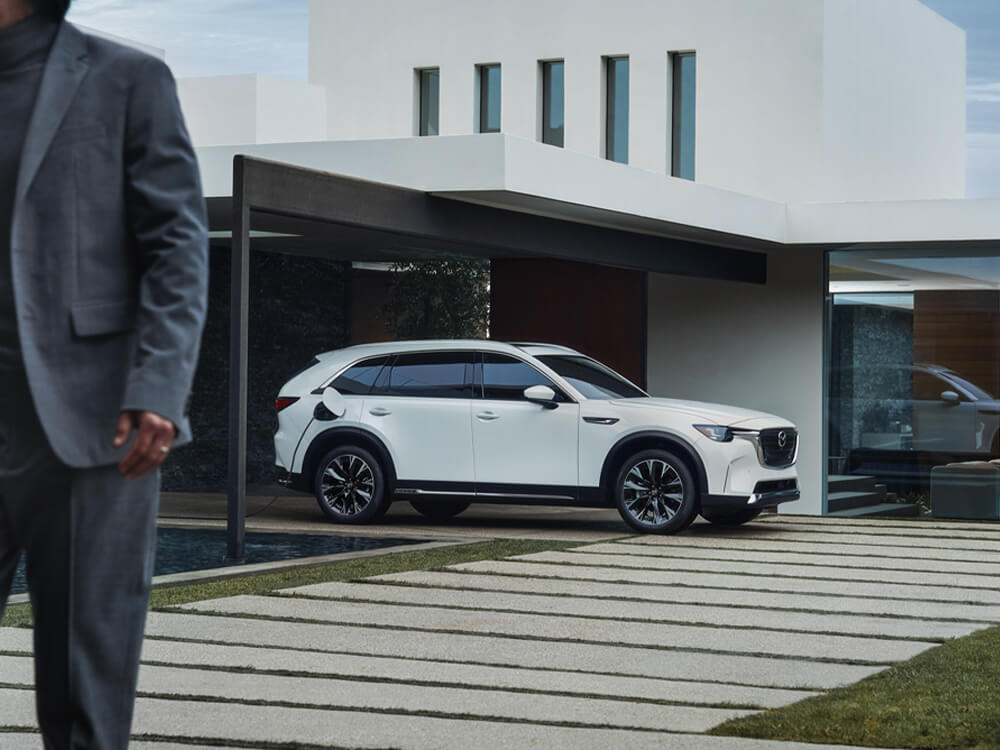 Plugged in and charging Arctic White CX-90 Plug-In Hybrid parked in front of stylish residence. A man in the close foreground is out of focus, walking away. 