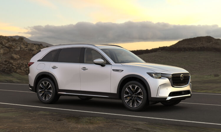 Close up of “CX-90 AWD” vehicle badge.  ¾ front shot of Arctic White CX-90 driving through hilly countryside at dawn/dusk.  