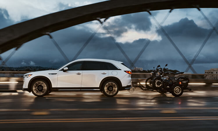 Arctic White CX-90 PHEV driving over a bridge at dusk, towing a small trailer with 2 motorcycles.  