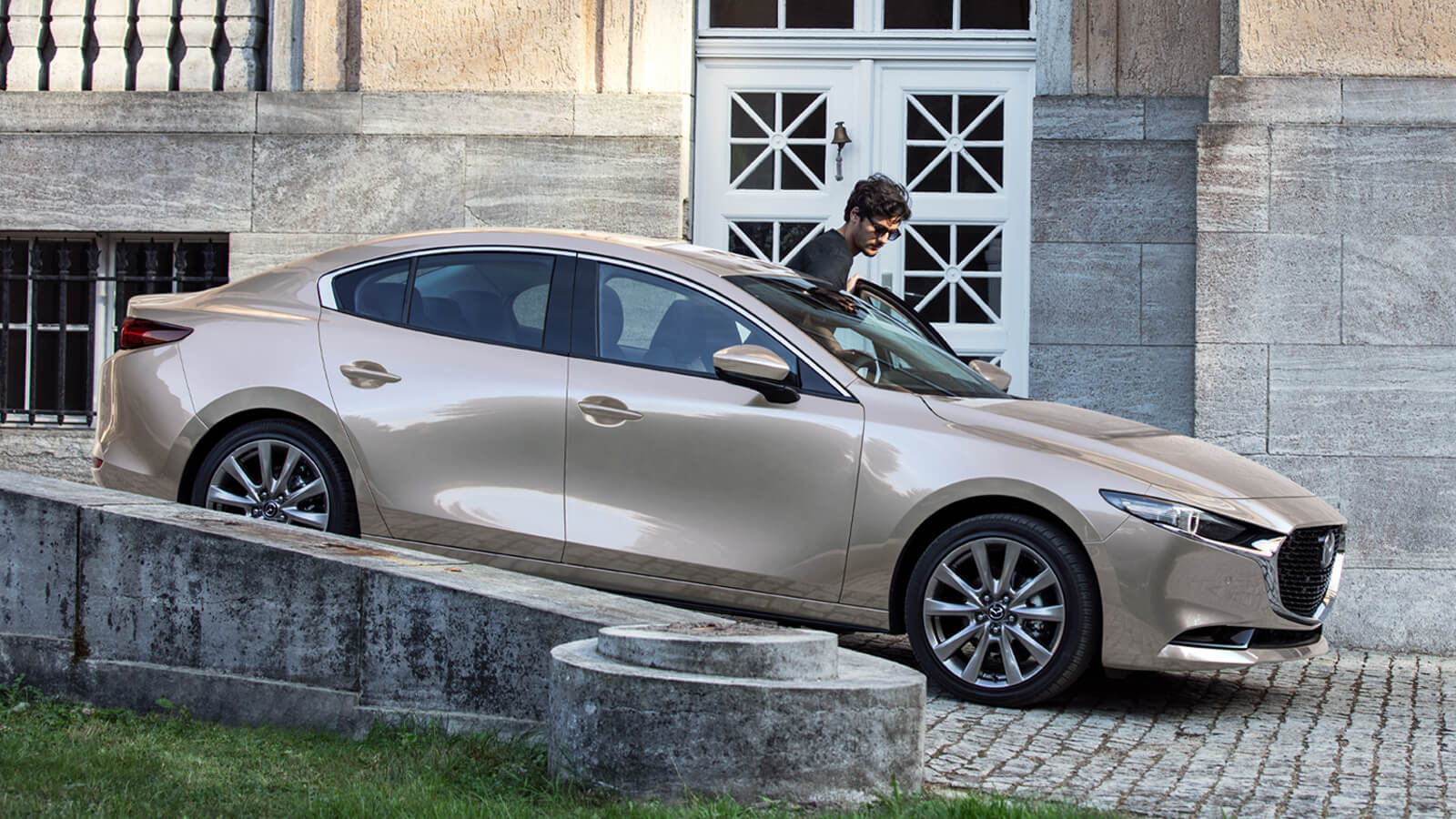 Driver stepping into a Mazda3 Sedan on cobblestone entryway in front of large limestone building. 