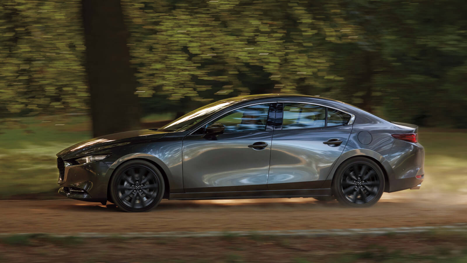 Mazda3 sedan drives swiftly along country road kicking up dust, passing a large tree in the background. 