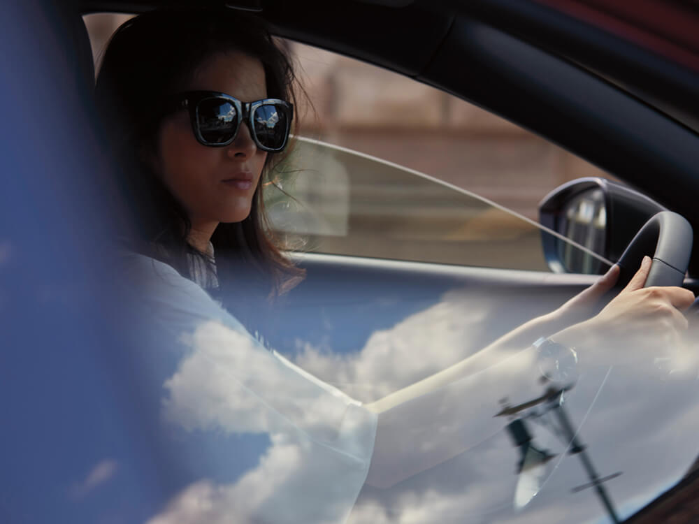 Front passenger window POV looking at female driver in Mazda3 sedan cockpit. Clouds and sky are reflected in the window. 