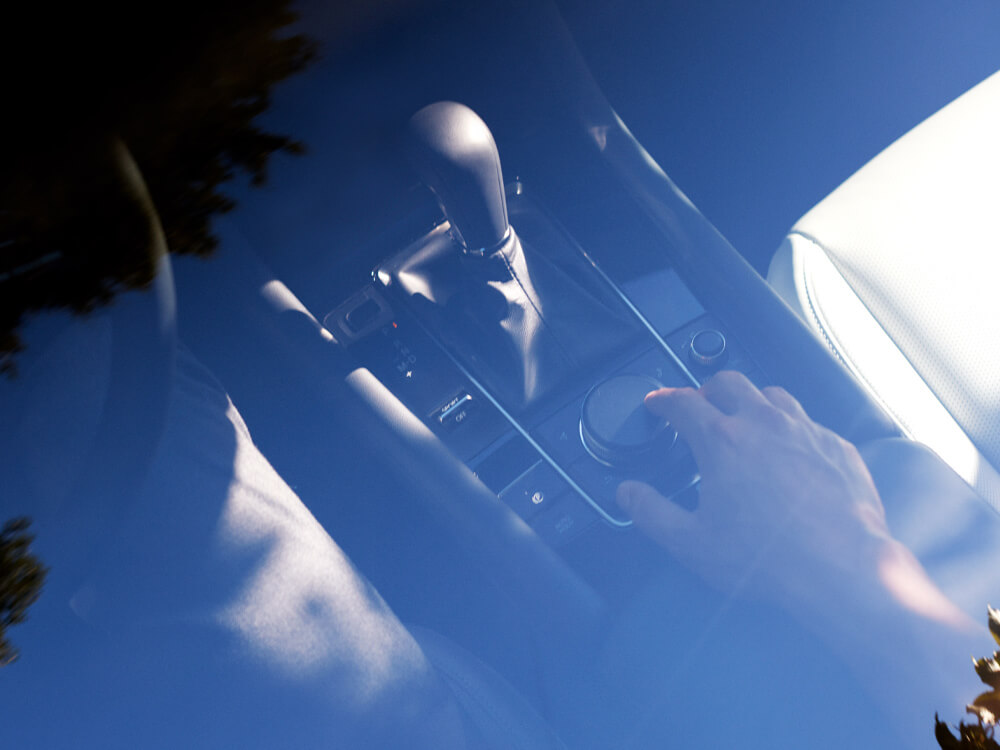 Looking down through closed moonroof, the driver is using the control knob on centre console of Mazda3 Sedan. 